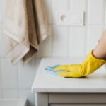 Mão com luva amarela a limpar bancada de lavatório com pano azul.