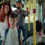 interior de autocarro com mulher, homem e criança no corredor entre as cadeiras. várias pessoas sentadas.