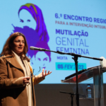 Presidente da CIG, Sandra Ribeiro, a falar no púlpito, com microfone. em fundo, a imagem do 6.º Encontro Regional para a Intervenção Integrada pelo Fim da Mutilação Genital Feminina