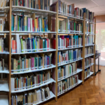 Sala com estantes preenchidas com livros. Planta de folhas verdes no canto inferiordireito da sala. Ao fundo, porta de acesso ao jardim.