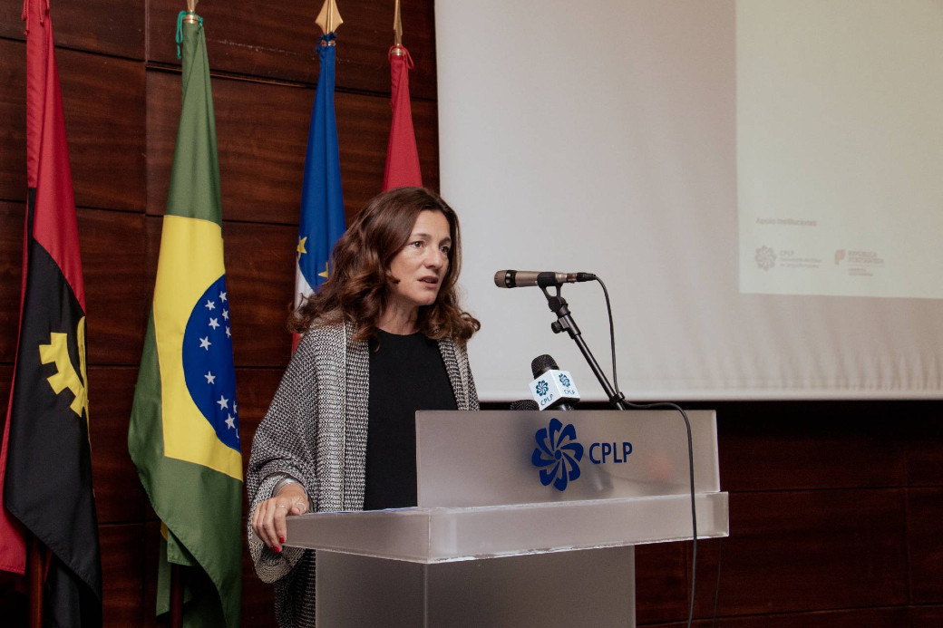 Presidnete da CIG, Sandra Ribeiro, fala num púplpito com o logótipo da CPLP. em fundo, bandeiras do Brasil, Cabo Verde e Angola