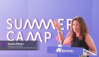Presidente da CIG, Sandra Ribeiro, fala num púlpito. Em fundo, parede lilás com referência Summer Camp