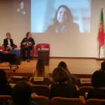 Painel com 3 mulheres no palco e uma projetada online. Bandeiras de Portugal, Noruega, Islândia e Liechtenstein . Interprete de LÍngua Gestual Portuguesa à esquerda