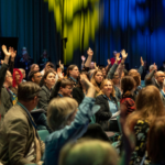 Homens e mulheres, participantes na conferência, com braços levantados solicitam intervenção