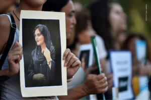 Fotografia de uma manifestação com os manifestantes desfocados a exibir um cartaz com a fotografia de Mahsa Amini