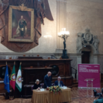 sala da assembleia da república. mesa ao centro com e homens e uma mulher. o homem do centro está de pé virado para a sua esquerda. em fundo, um enorme quadro na parede. ao lado da mesa, um estandarte lilás e letras brancas Acom a inscrição prémio direitos humanos da assembleia da república