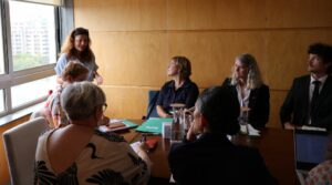 Fotografia da intervenção da Presidente da CIG, que se encontra em pé e os restantes elementos sentados à mesa de reuniões