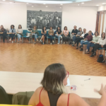 Fotografia da sessão de formação, a oradora surge no centro de costas e as pessoas formandas sentadas em secretárias em formato de u