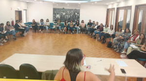 Fotografia da sessão de formação, a oradora surge no centro de costas e as pessoas formandas sentadas em secretárias em formato de u 